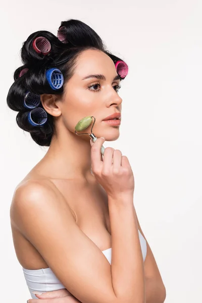 Jovem mulher em curlers e top fazendo massagem facial com rolo de jade isolado em branco — Fotografia de Stock
