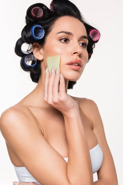 Grave jovem mulher em curlers e top fazendo massagem facial com gua sha isolado em branco — Fotografia de Stock