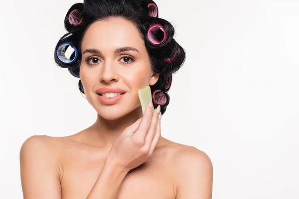 Smiling young woman in curlers and top making facial massage with gua sha isolated on white — Stock Photo