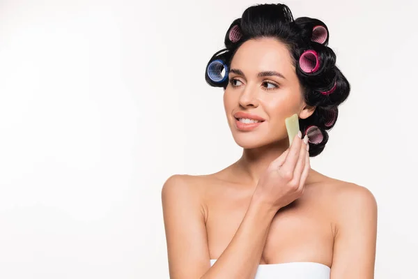 Young woman in curlers and bra making facial massage with gua sha scraper isolated on white — Stock Photo