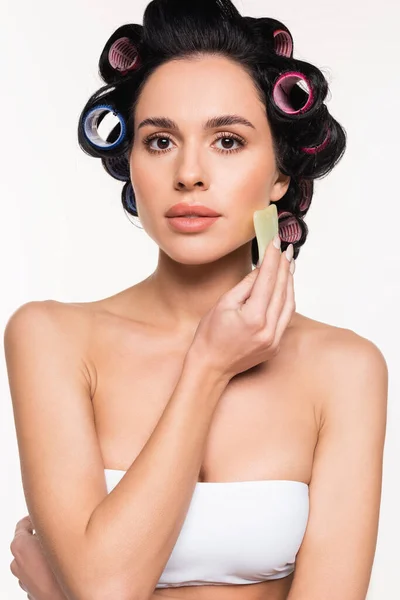 Seria giovane donna in bigodini e top fare massaggio facciale con gua sha raschietto isolato su bianco — Foto stock