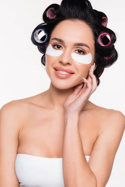 Smiling young woman in curlers with eye patches holding hand near face isolated on white — Stock Photo