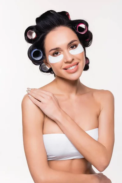 Smiling young woman in curlers and bra with eye patches on face holding hand on shoulder isolated on white — Stock Photo