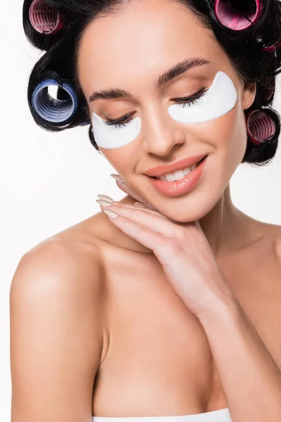 Retrato de mujer joven sonriente en rulos con parches en los ojos sosteniendo la mano cerca de la cara con los ojos cerrados aislados en blanco - foto de stock