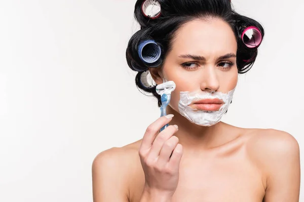 Imbarazzato giovane donna in bigodini tenendo rasoio vicino al viso con crema da barba isolato su bianco — Foto stock