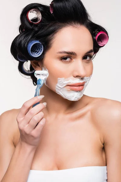 Jovem mulher séria em rolos segurando navalha perto do rosto com creme de barbear isolado no branco — Fotografia de Stock