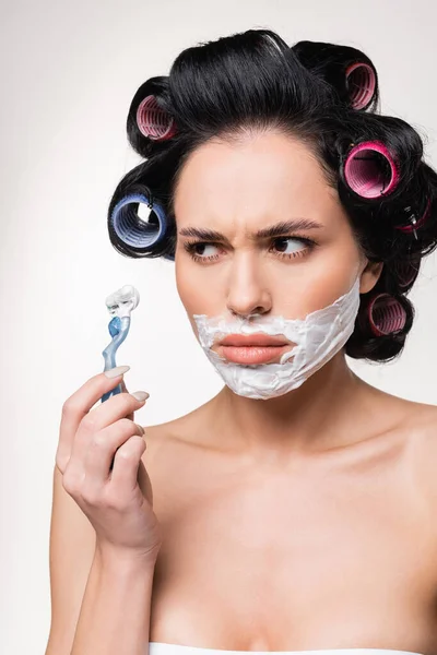 Mujer joven confundida en rulos sosteniendo afeitadora cerca de la cara con crema de afeitar aislado en blanco - foto de stock