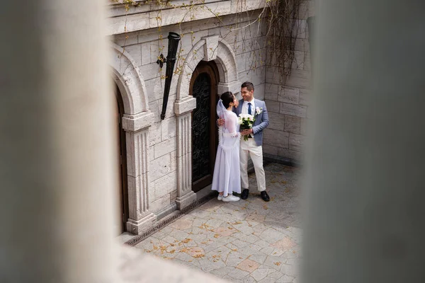 Cheerful groom embracing young wife with bouquet near building — Stock Photo