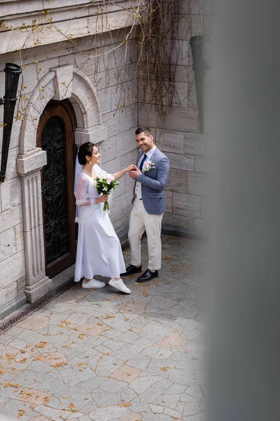 Groom holding hand of smiling bride with bouquet and smiling at camera near building — Stock Photo