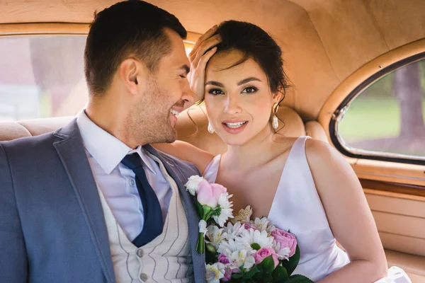 Positive bride with bouquet looking at camera near groom in retro car — Stock Photo