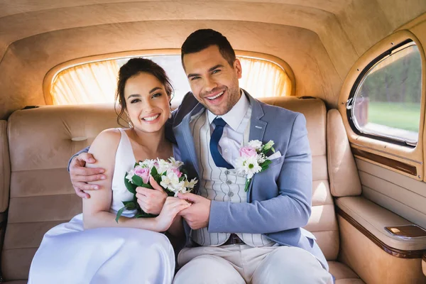 Happy groom embracing bride with bouquet in auto — Stock Photo