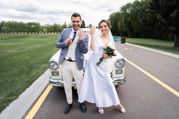 Alegre recién casados mostrando anillos cerca de auto retro en la carretera - foto de stock