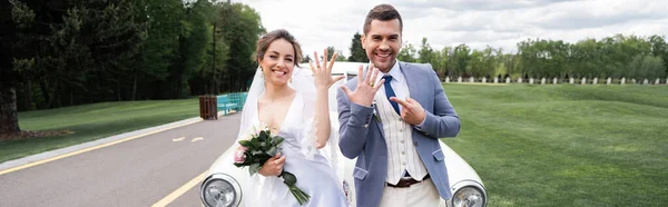 Веселый жених указывая на кольцо рядом с невестой с букет и винтажный автомобиль, баннер — стоковое фото