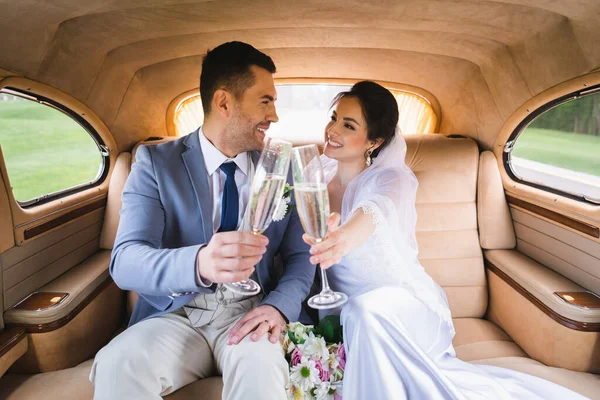 Sonrientes recién casados mirándose mientras brindan con champán en el coche - foto de stock