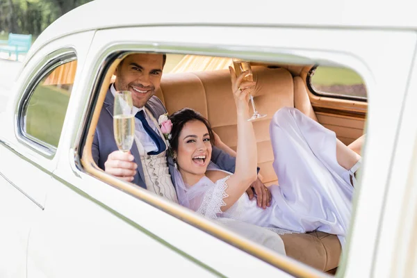 Cheerful groom holding blurred glass of champagne near bride in retro car — Stock Photo