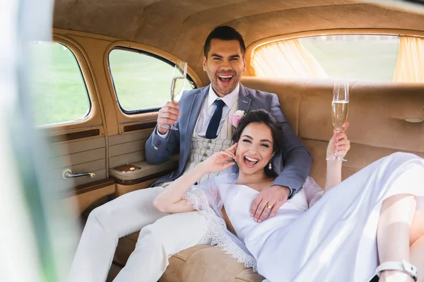 Happy newlyweds with glasses of champagne looking at camera in car — Stock Photo