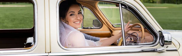 Улыбающаяся невеста смотрит в камеру на водительском сиденье винтажного автомобиля, баннер — стоковое фото