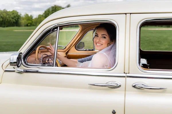 Bride in veil smiling at camera on driver seat of retro car — Stock Photo