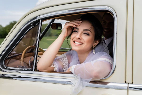 Счастливая невеста смотрит в камеру возле окна винтажного авто — стоковое фото