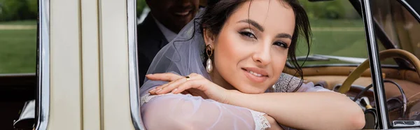 Novia sonriente en velo mirando a la cámara cerca del novio borroso en el coche vintage, pancarta - foto de stock