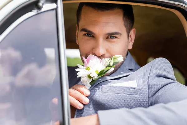 Elegante novio oliendo flores de boutonniere en coche retro - foto de stock