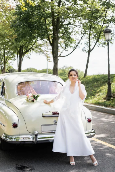 Mariée souriante regardant la caméra près de voiture vintage sur la route — Photo de stock