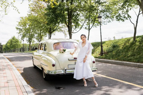 Mariée souriante en robe et voile debout près de voiture rétro — Photo de stock