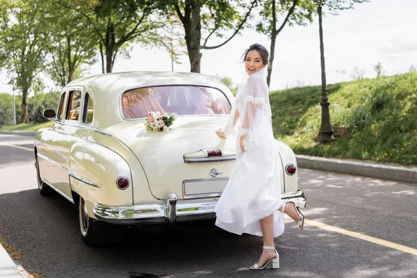Mariée heureuse debout près du bouquet sur la voiture vintage sur la route — Photo de stock