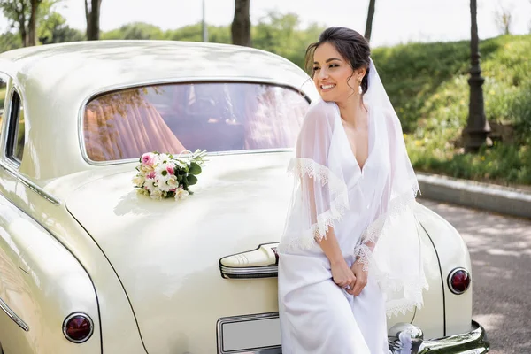 Noiva feliz em vestido de noiva de pé perto do buquê em retro auto — Fotografia de Stock