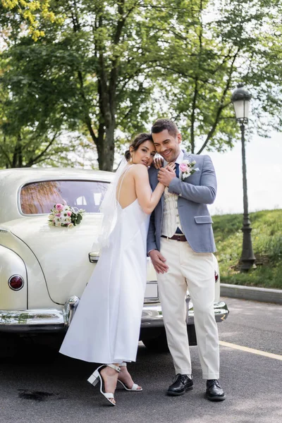Femme heureuse en robe de mariée embrassant marié près de la voiture sur la route — Photo de stock