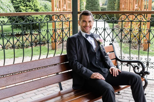 Cheerful groom in suit looking at camera on bench in patio — Stock Photo