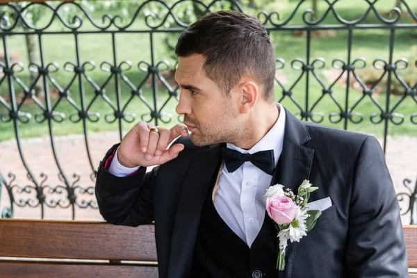 Vue latérale du marié avec boutonnière sur veste regardant loin sur banc — Photo de stock