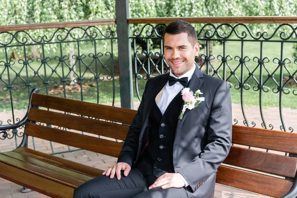 Sorrindo noivo com boutonniere olhando para a câmera no banco no pátio — Fotografia de Stock