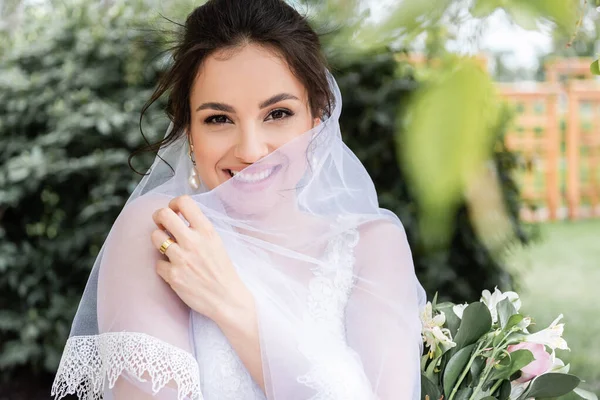 Cheerful bride holding white veil near bouquet outdoors — Stock Photo