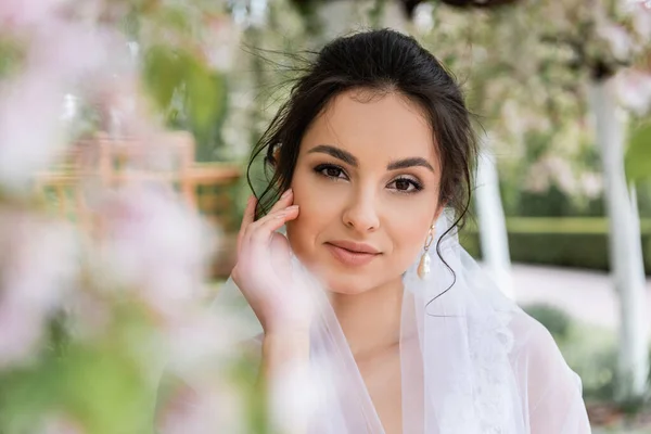 Hübsche Braut mit weißem Schleier blickt im Park in die Kamera — Stockfoto