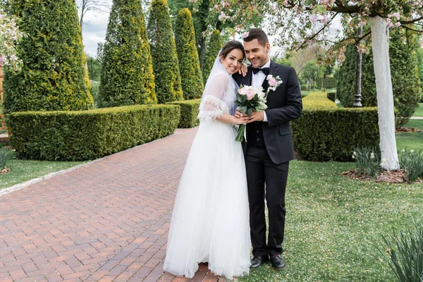 Cheerful bride with bouquet hugging groom in park — Stock Photo