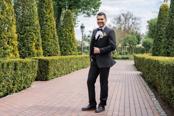 Smiling groom in suit with boutonniere standing in park — Stock Photo