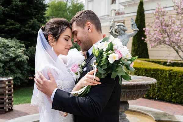 Vue latérale du marié souriant embrassant mariée avec bouquet près fontaine floue — Photo de stock