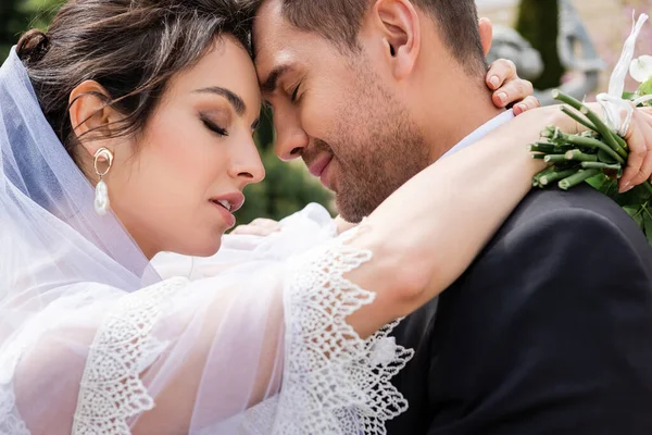 Vista lateral de la novia con los ojos cerrados abrazando novio en traje al aire libre - foto de stock