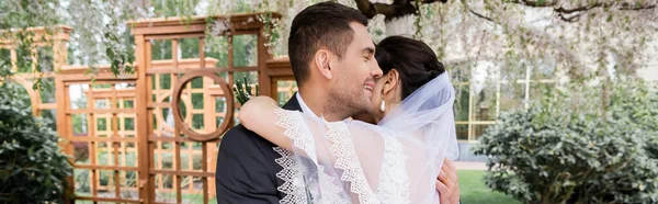 Giovane sposo sorridente che abbraccia sposa in velo nel parco, banner — Foto stock