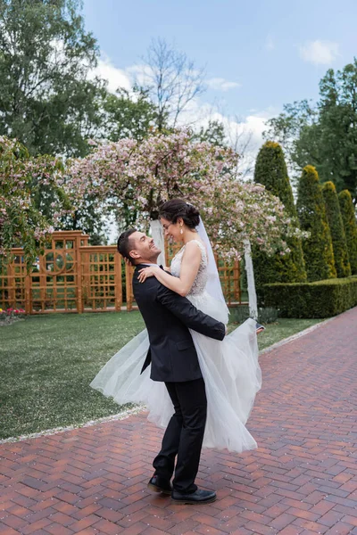 Vista laterale di sposo sorridente che tiene su mani la sposa carina in parco — Foto stock