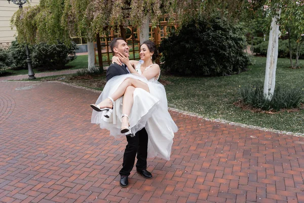 Novia alegre en velo y vestido de novia tocando la cara del novio en el parque - foto de stock