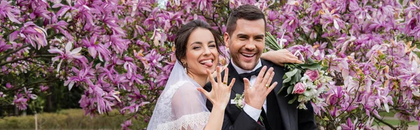 Cheerful bride and groom showing rings and looking at camera near magnolia trees, banner — Stock Photo