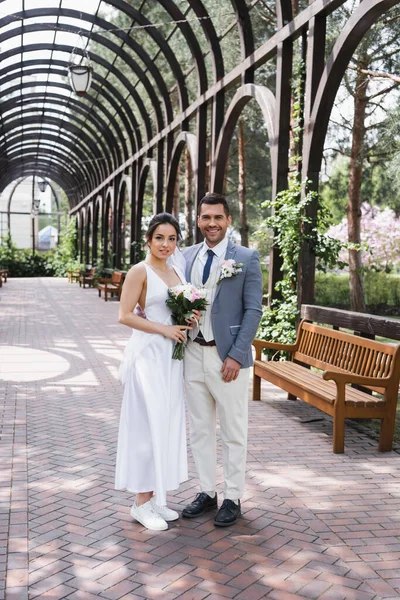 Sonriente novia mirando a la cámara cerca del novio en el parque - foto de stock