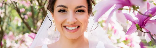 Junge brünette Braut mit Schleier blickt in die Kamera in der Nähe blühender Magnolien, Banner — Stockfoto