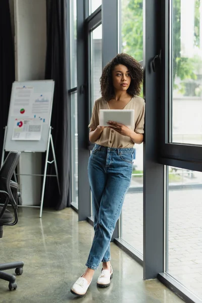 Thoughtful african american woman in casual clothes standing near window with digital tablet in hand in modern office — Stock Photo