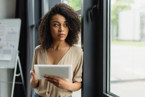 Mujer afroamericana reflexiva de pie cerca de la ventana con la tableta digital en la mano en la oficina moderna - foto de stock