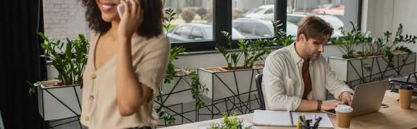 Junger Mann, der mit Laptop in der Nähe eines afrikanisch-amerikanischen Kollegen arbeitet, der im modernen Büro mit dem Handy spricht, Banner — Stockfoto