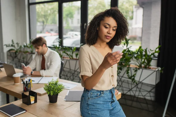 Молодая африканская американка, обменивающаяся сообщениями по мобильному телефону рядом с размытым коллегой, сидящим рядом с ноутбуком в современном офисе — стоковое фото