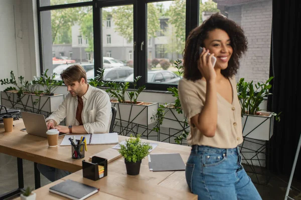 Giovane che lavora con un computer portatile vicino a un collega afroamericano offuscato che parla al cellulare in un ufficio moderno — Foto stock